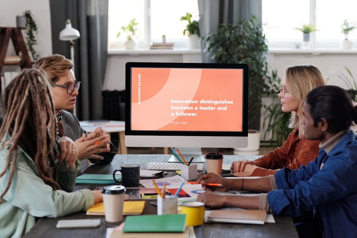 Attractiv Movies A multicultural team collaborating in an office, discussing a project with a visible monitor.